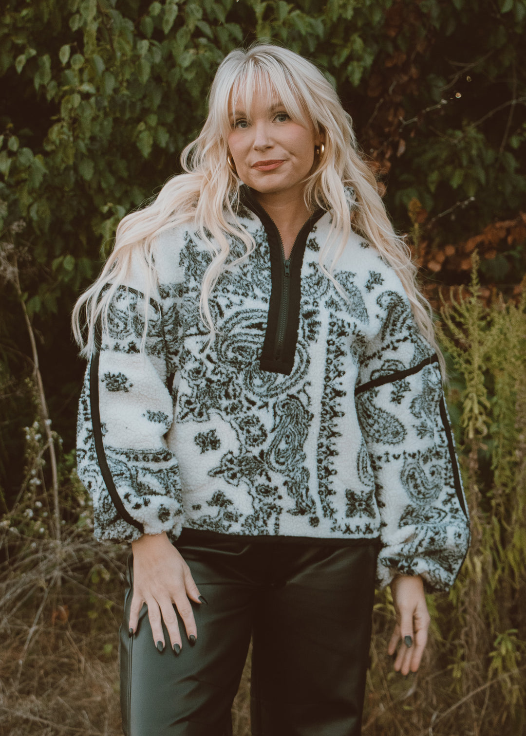 Woman wearing a patterned jacket outdoors with greenery in the background