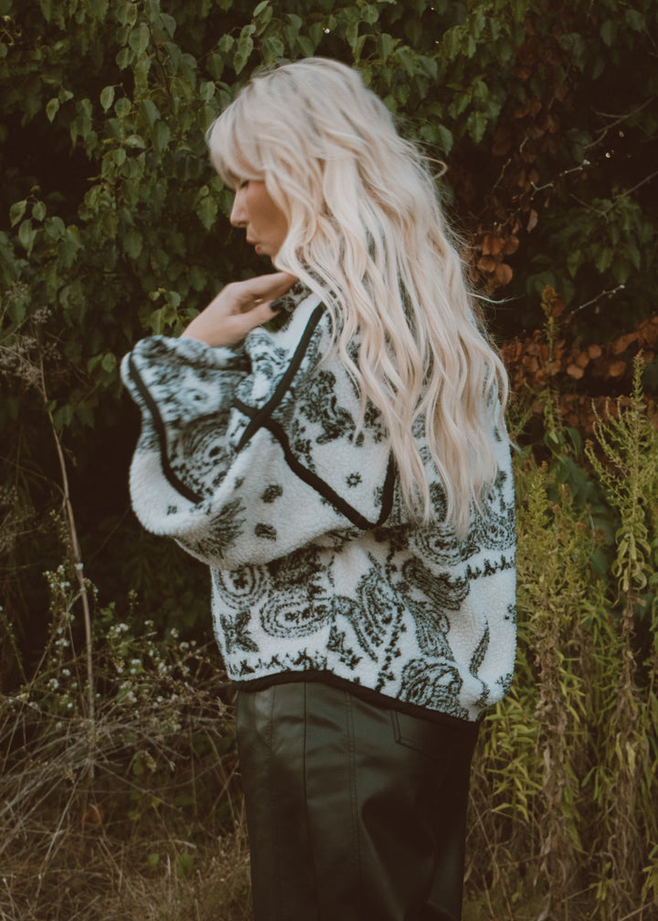 Person wearing a patterned sweater jacket standing in a natural setting with greenery.