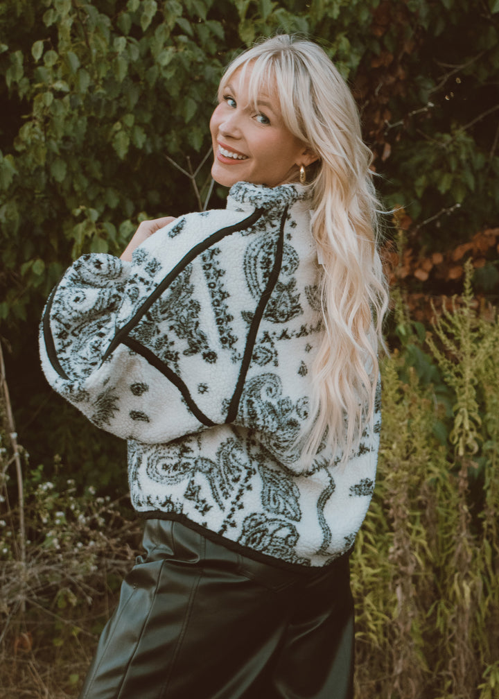 Woman wearing a patterned sweater standing outdoors with greenery in the background