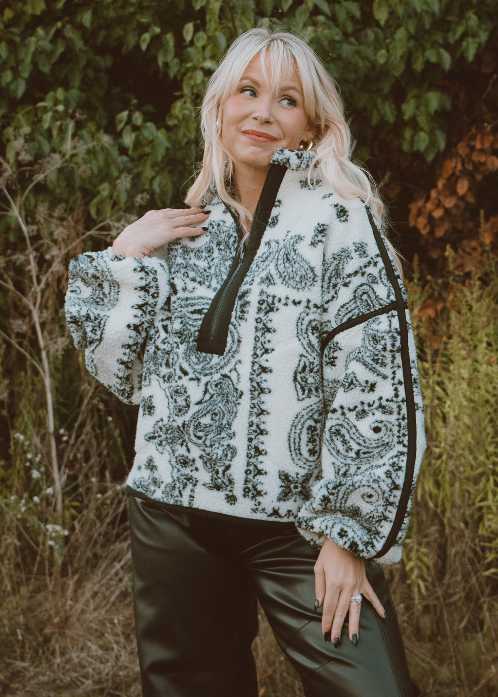 Woman wearing a patterned jacket standing outdoors with greenery in the background
