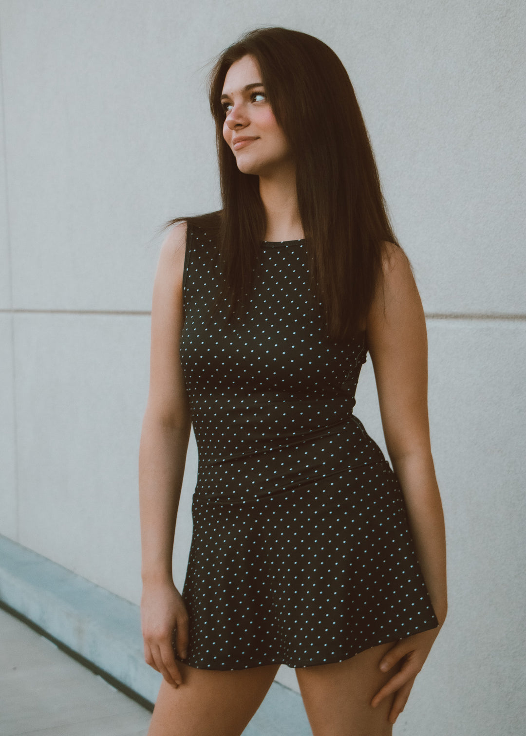 Woman wearing a black polka dot dress against a light gray wall.
