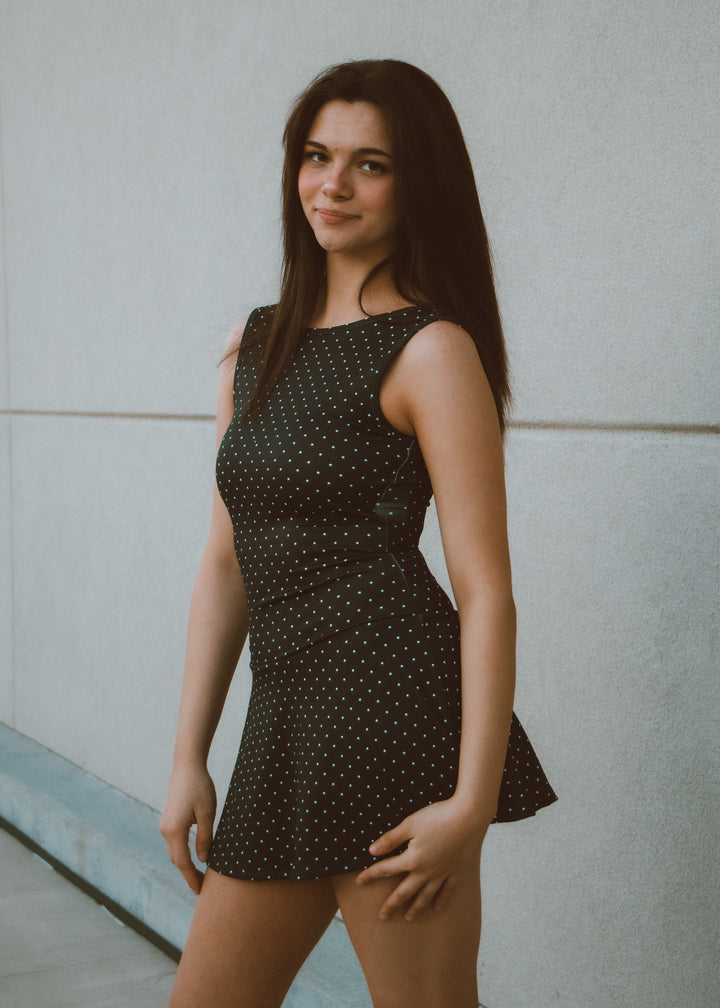 Woman wearing a black polka dot dress against a light gray wall.