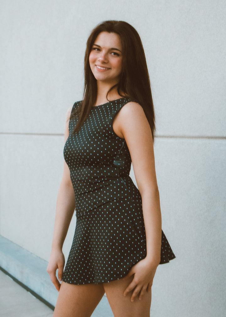 Woman wearing a black polka dot dress standing against a light-colored wall.