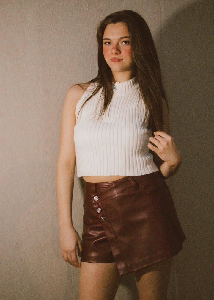 Woman wearing a white ribbed top and brown leather skirt against a plain background