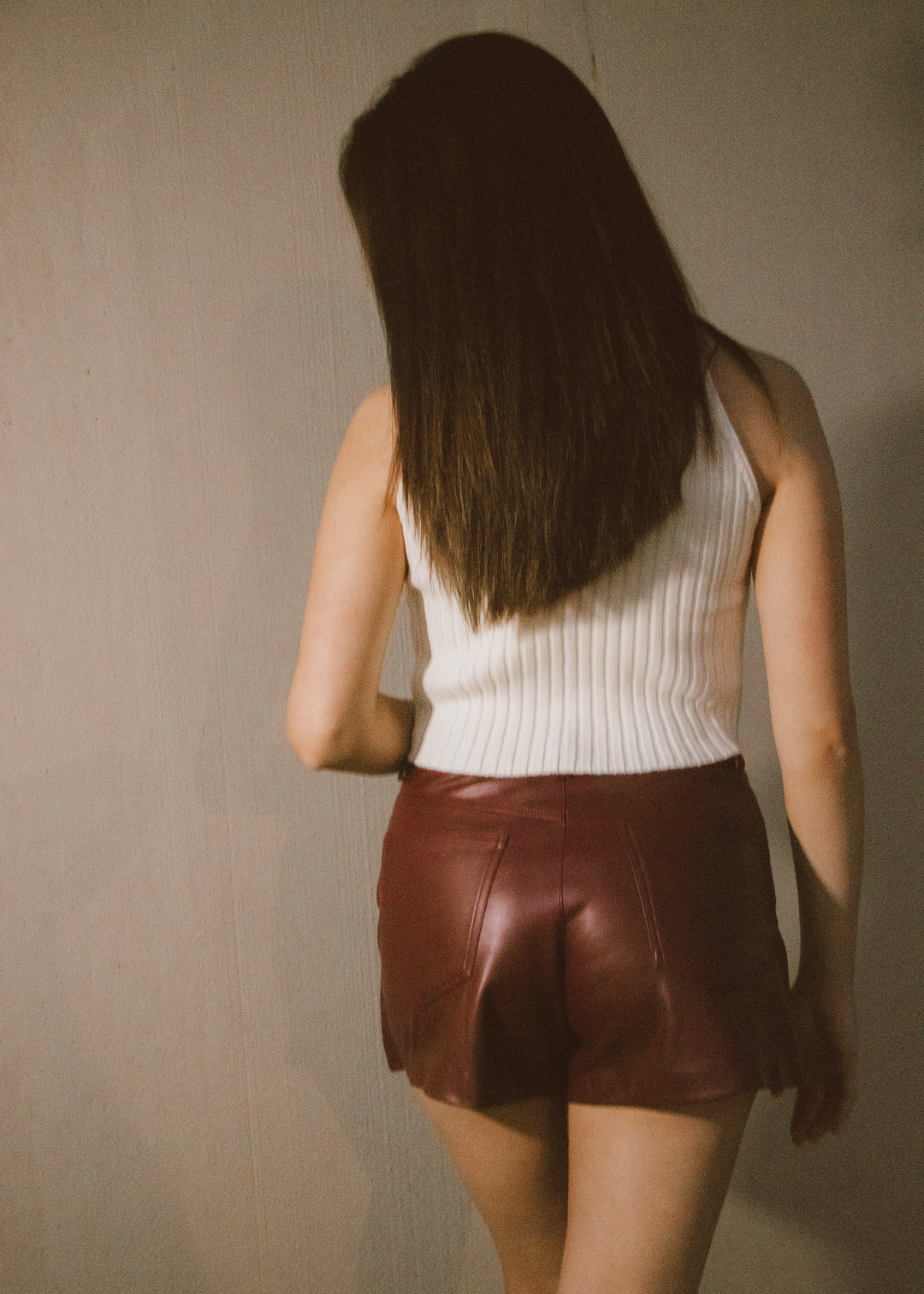 Person wearing a white sleeveless top and brown leather skirt against a plain wall.
