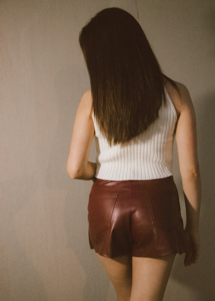 Person wearing a white sleeveless top and brown leather skirt against a plain wall.