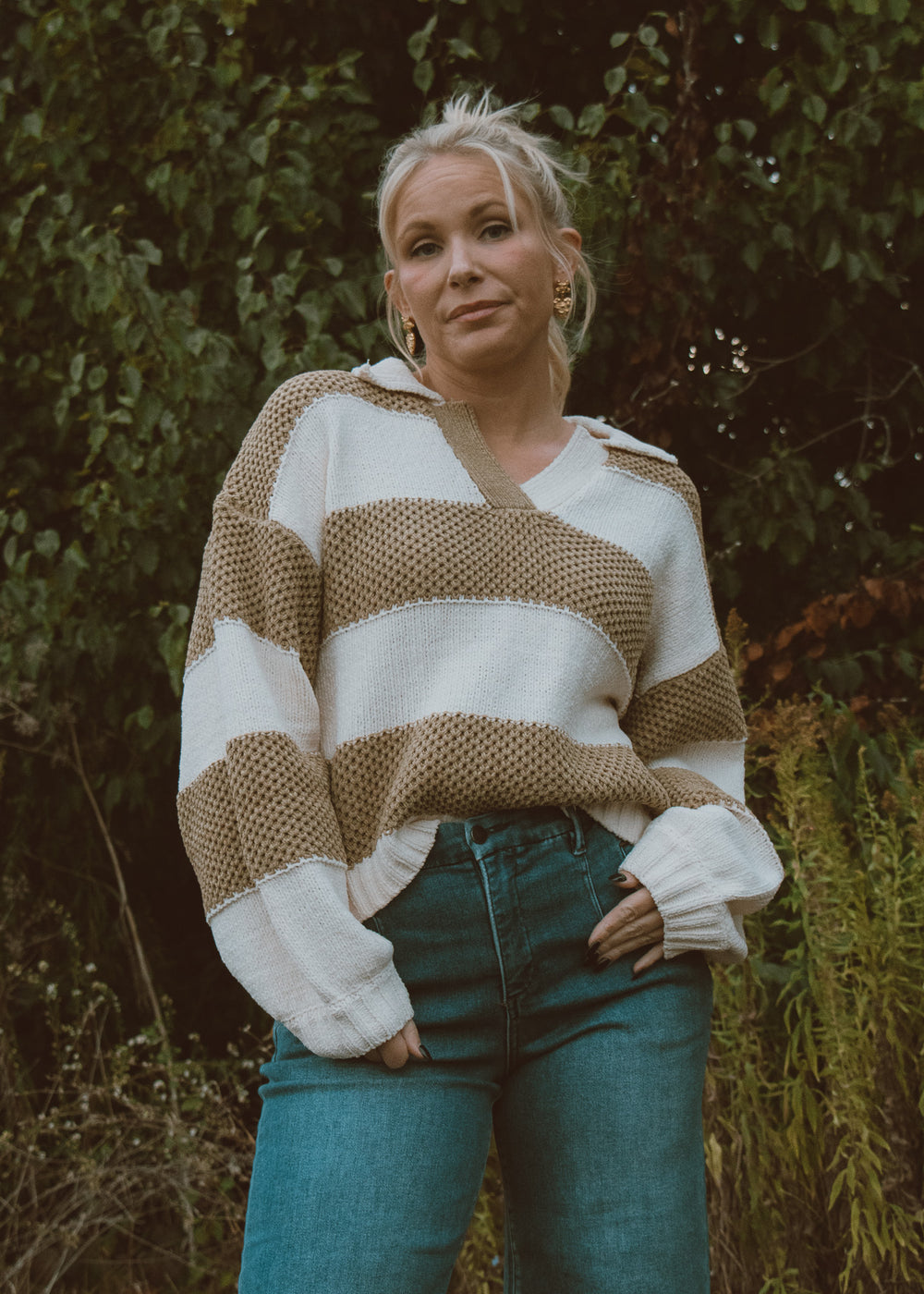 Woman wearing a green and white striped sweater with blue jeans standing in a natural setting.
