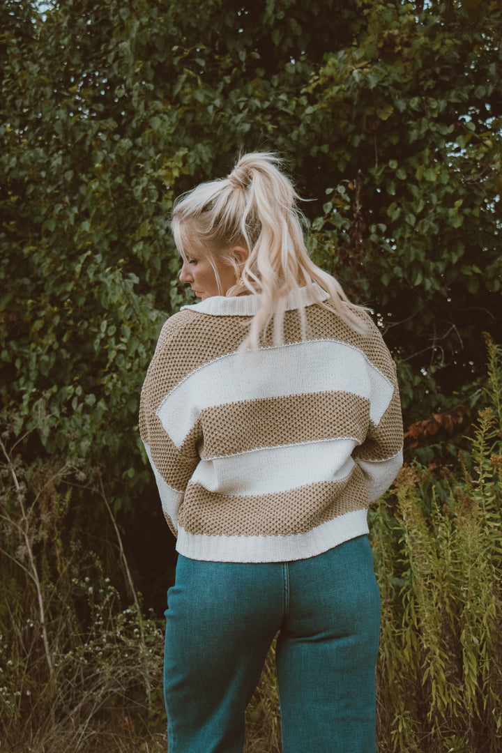 Woman wearing a striped sweater and blue jeans standing in a natural setting with trees.