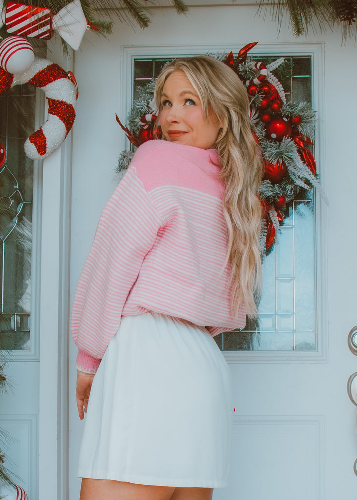 Woman wearing a pink sweater and white skirt standing in front of a decorated door with Christmas wreath and candy canes.