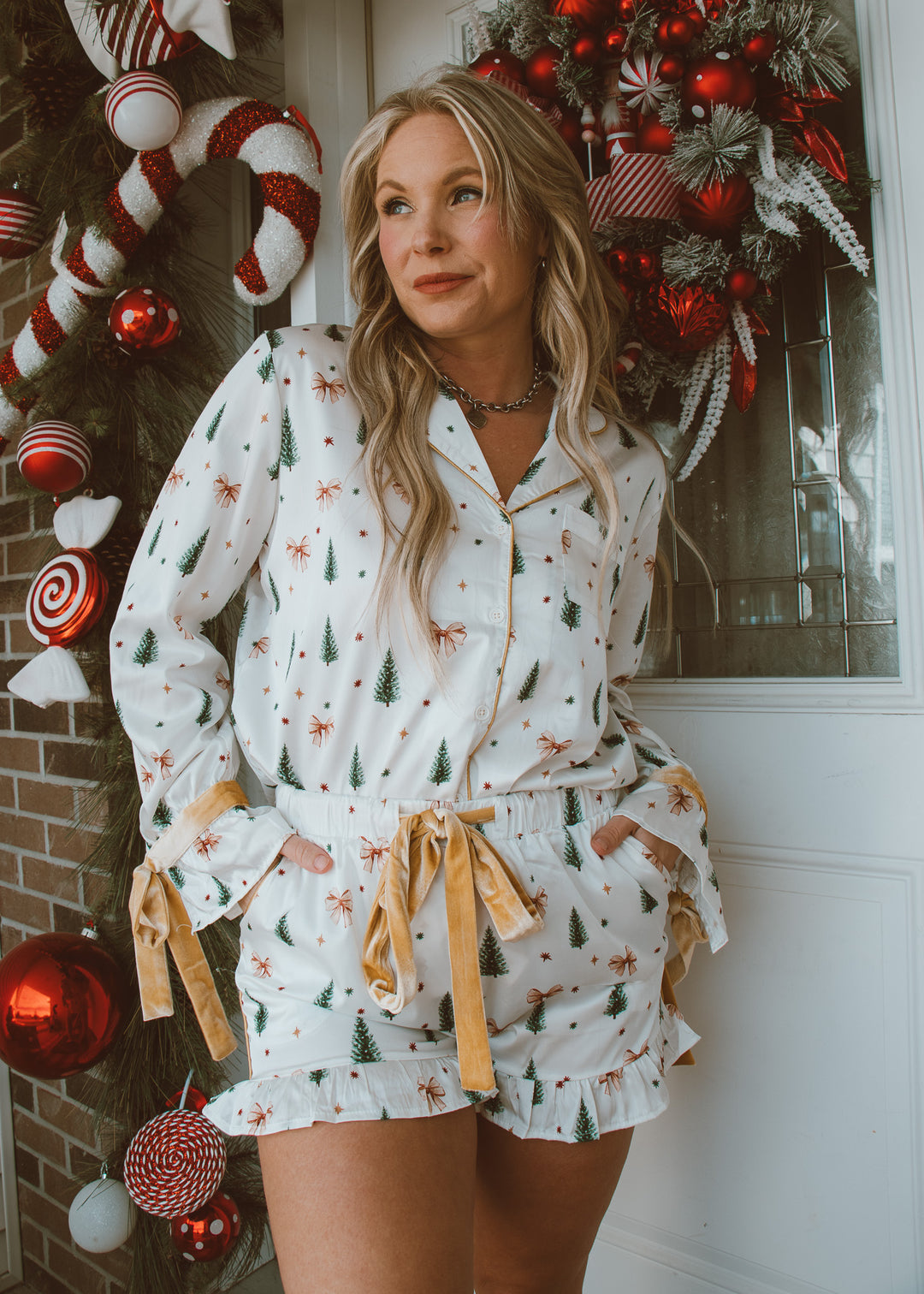 Woman wearing a festive pajama set with pine trees and bows in front of a decorated Christmas door.