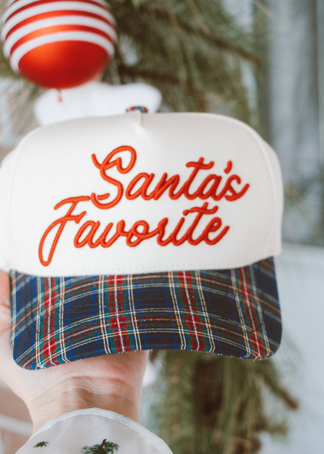 Cap with 'Santa's Favorite' text and plaid pattern, held against a blurred natural background.