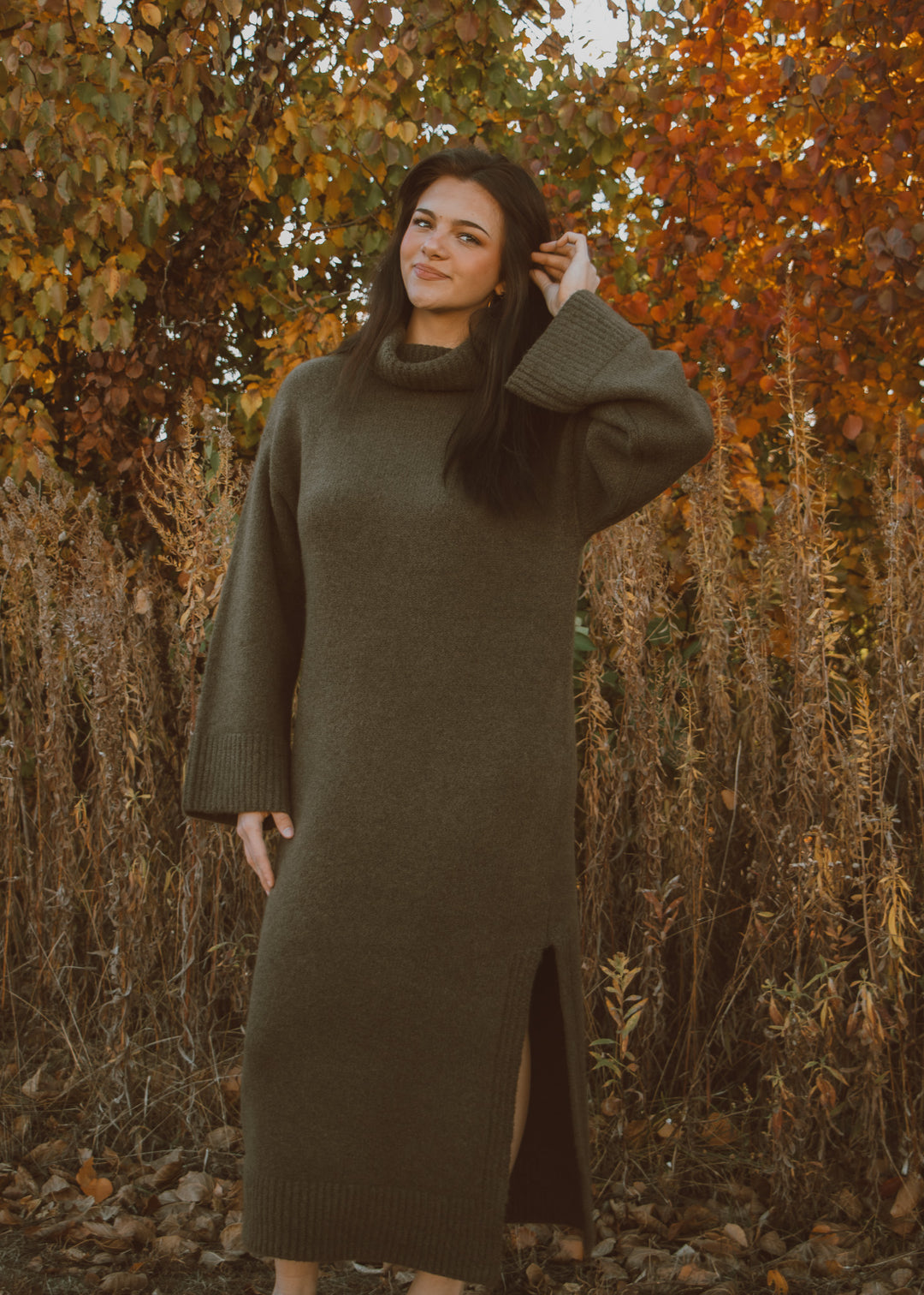 Woman wearing a green sweater dress standing in an autumnal setting with trees and dry grass.