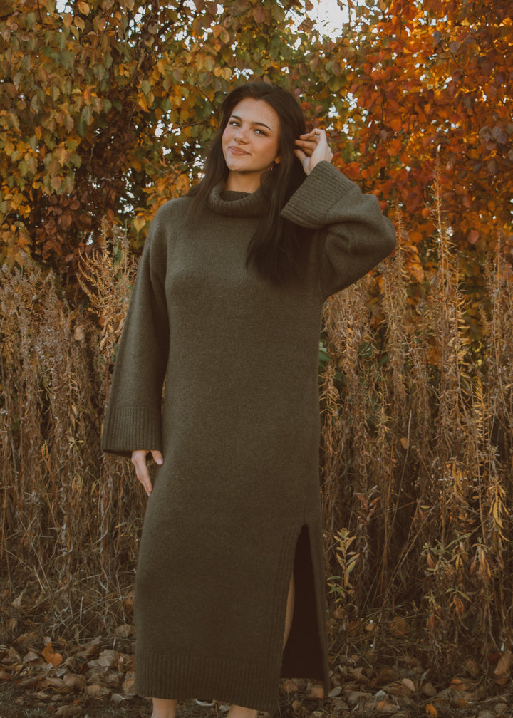 Woman wearing a green sweater dress standing in an autumnal setting with trees and dry grass.