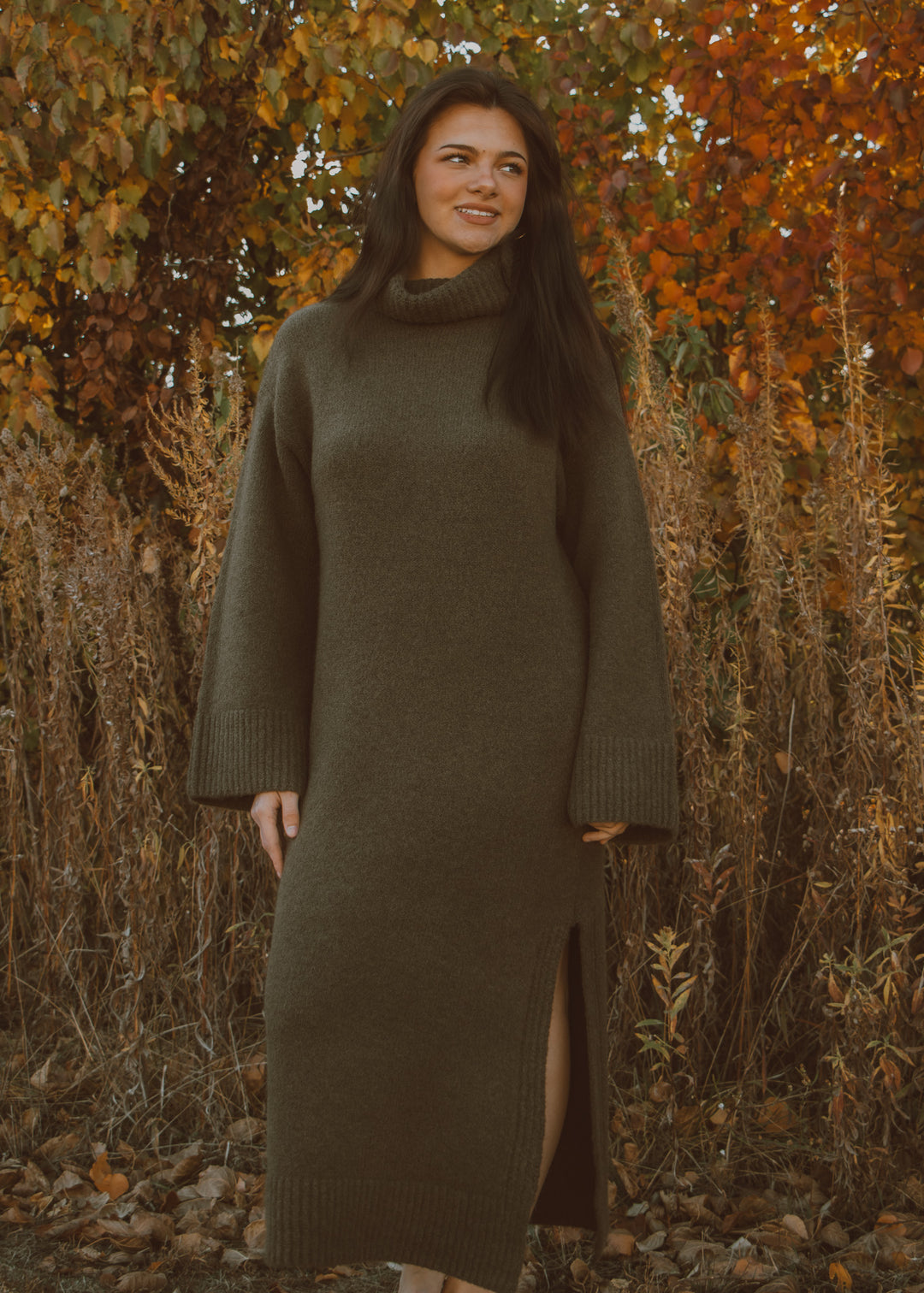 Woman wearing a long olive green sweater dress standing in an autumn forest.