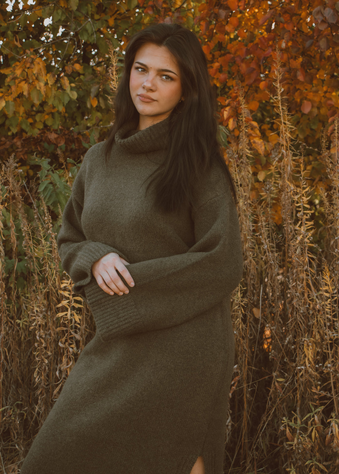 Woman wearing a green sweater dress standing in an autumnal setting with trees and dry grass.