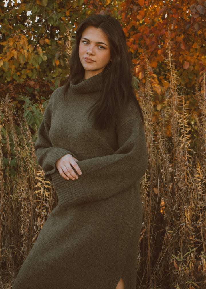 Woman wearing a green sweater dress standing in an autumnal setting with trees and dry grass.