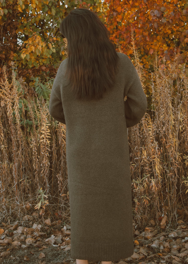 Person wearing a long green sweater dress standing in an autumnal setting with trees and dry grass.
