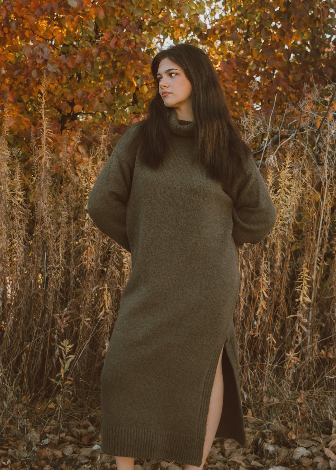 Woman wearing a long olive green sweater dress standing in an autumn forest.