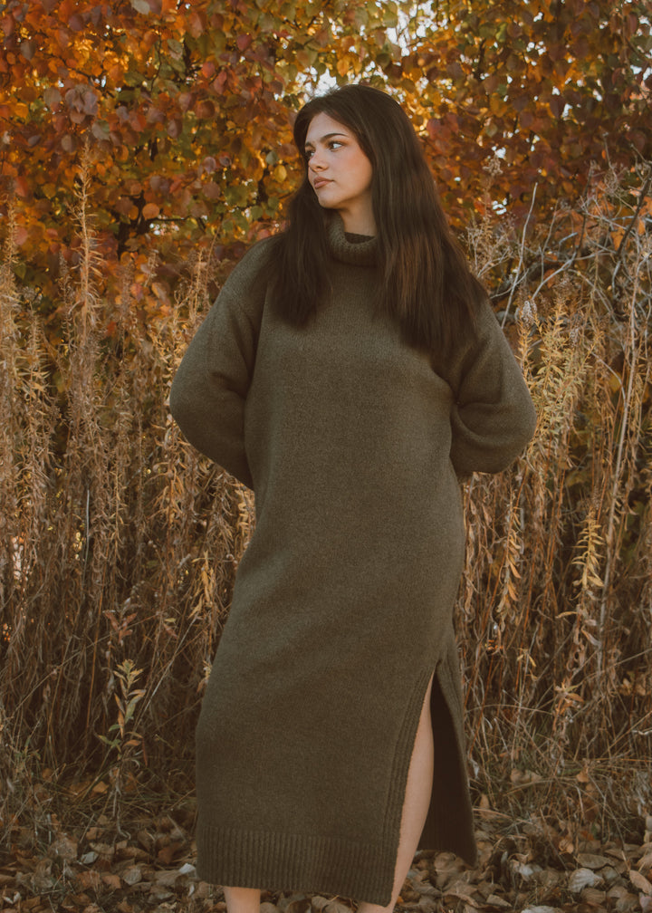 Woman wearing a long olive green sweater dress standing in an autumn forest.