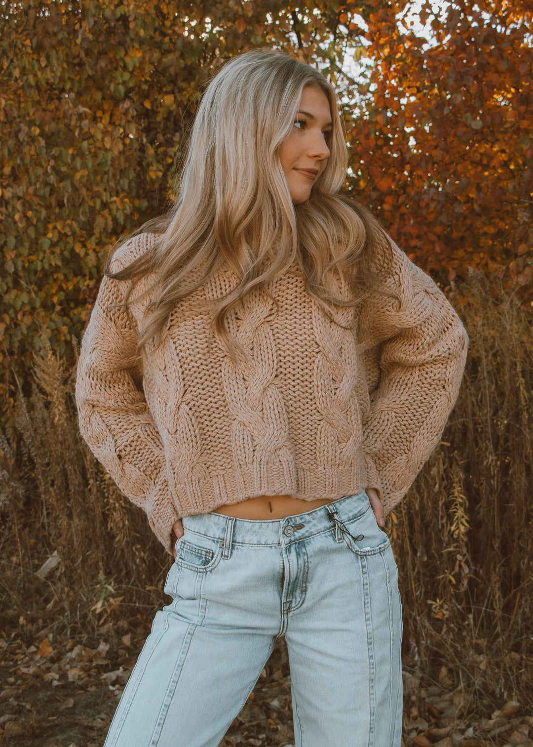 Woman wearing a tan cable knit sweater and light blue jeans standing in an autumn setting.