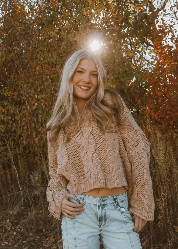Woman in a beige knitted sweater and light blue jeans standing in an autumn forest.