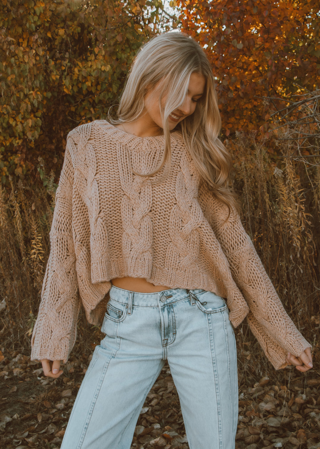 Woman wearing a beige knitted sweater and light blue jeans standing in an autumn setting.