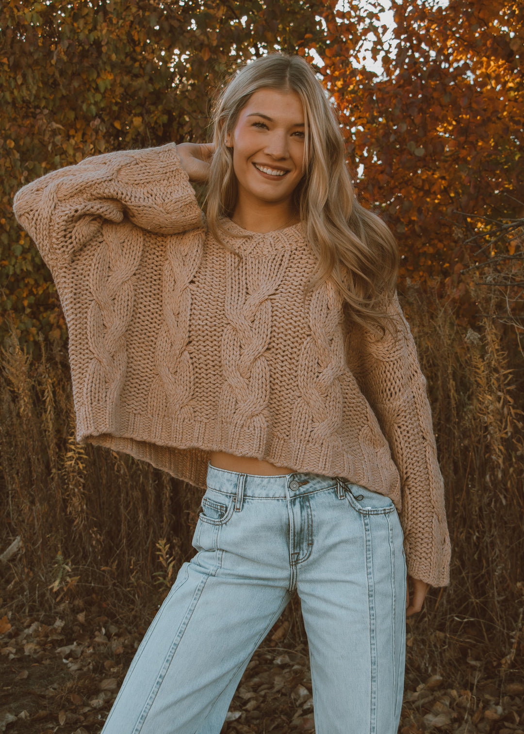 Woman wearing a beige cable knit sweater and light blue jeans standing in an autumn setting.