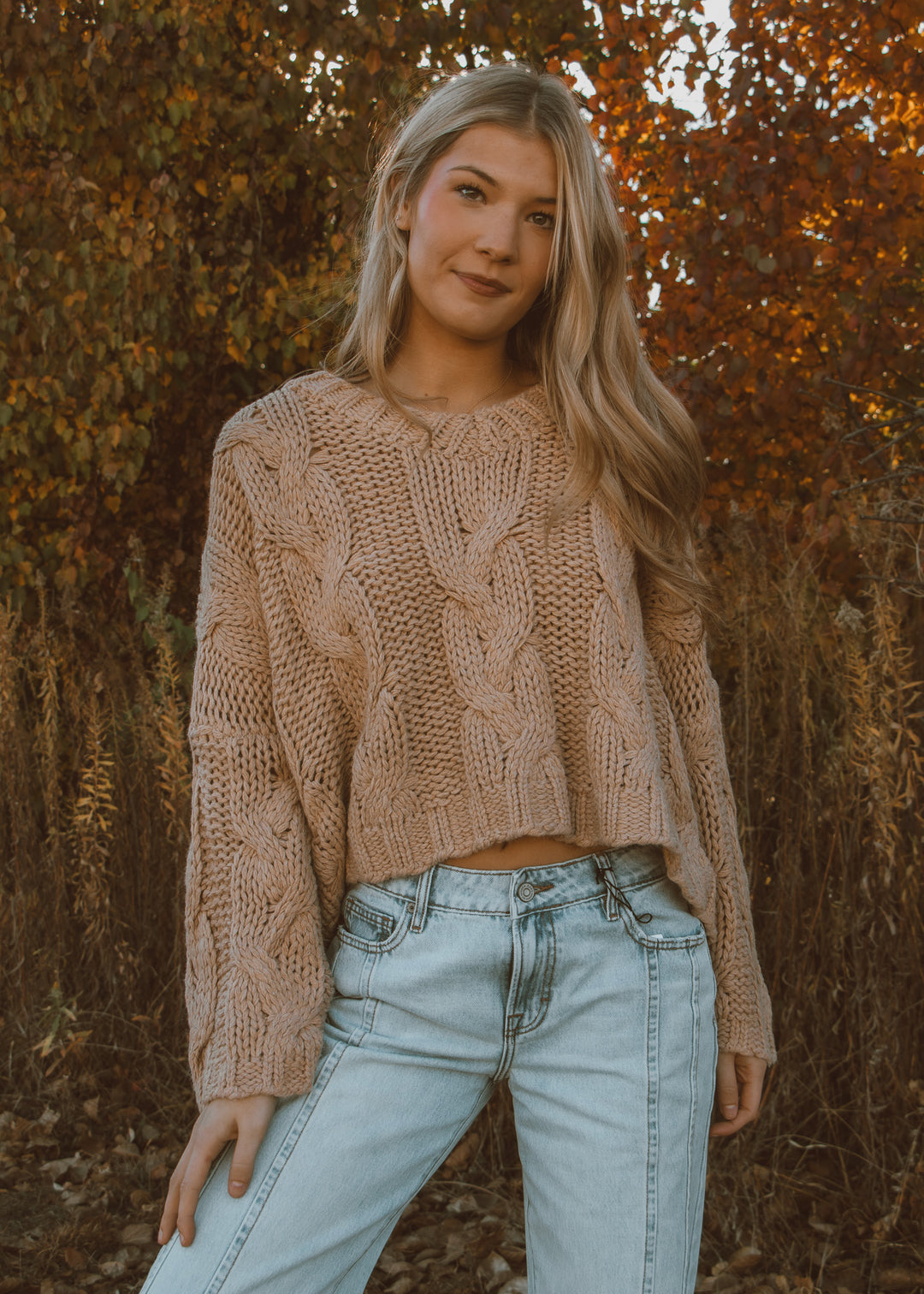 Woman wearing a tan sweater and light blue jeans standing in front of autumn foliage.