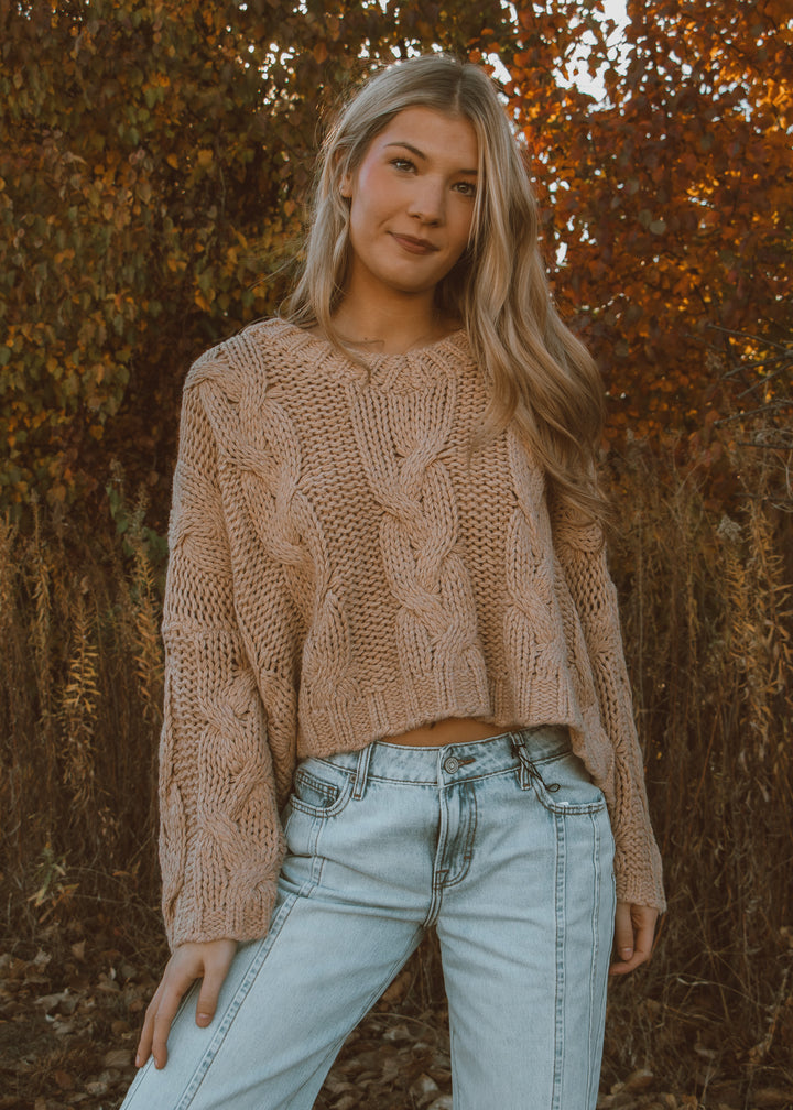 Woman wearing a tan sweater and light blue jeans standing in front of autumn foliage.