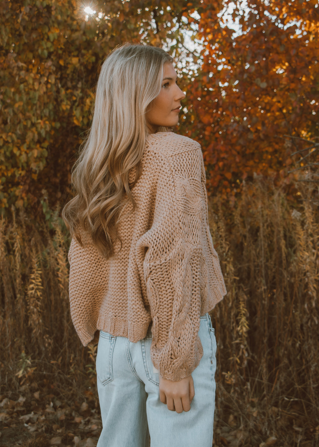 Woman wearing a tan sweater and light blue jeans standing in front of autumn foliage.