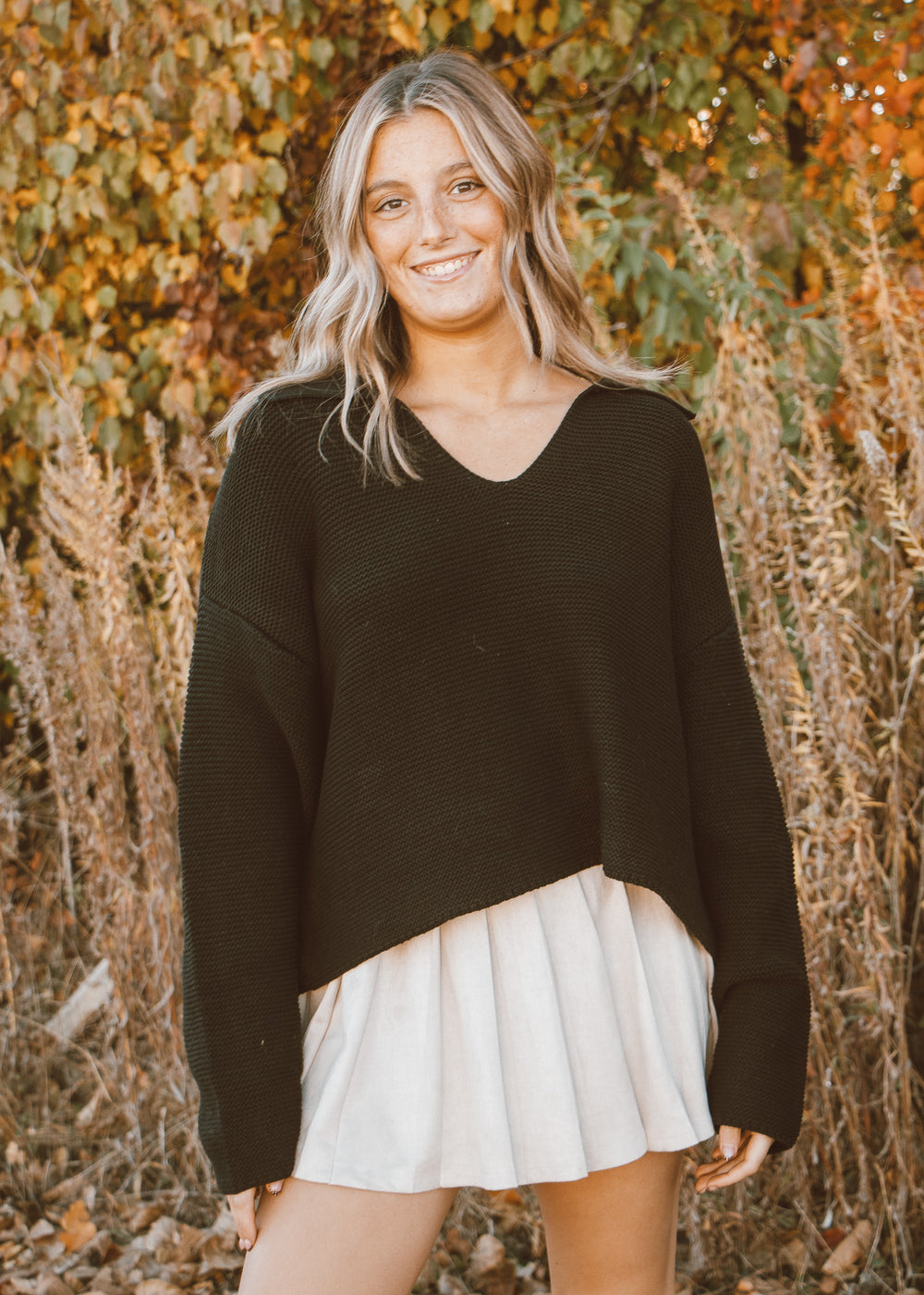 Woman wearing a black sweater and white skirt standing in front of autumn foliage.
