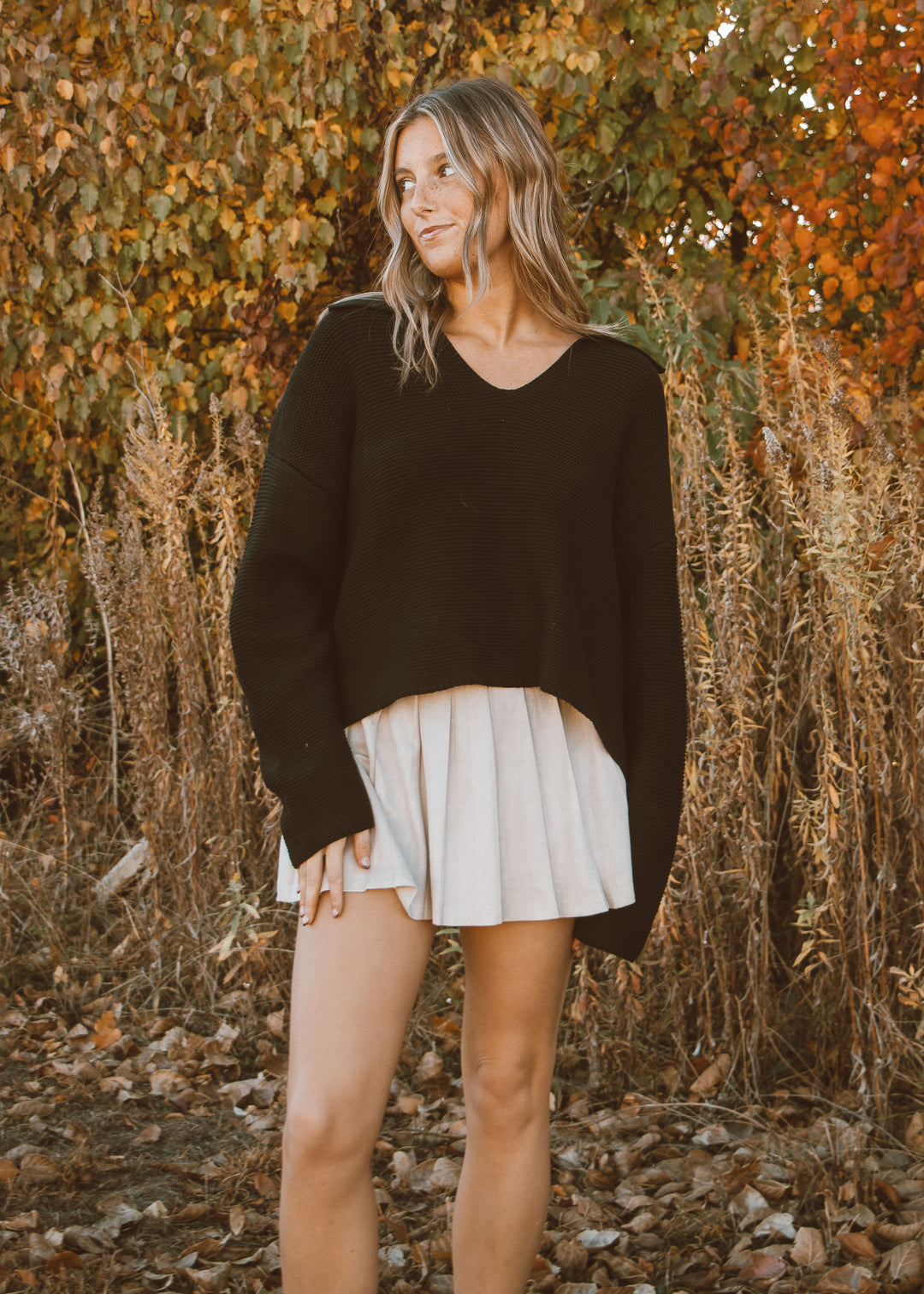 Woman wearing a black sweater and white skirt standing in front of autumn foliage.
