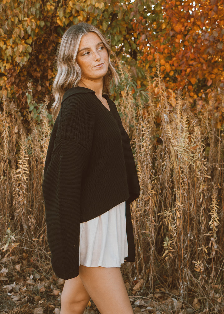 Woman in a black sweater and white skirt standing in an autumn setting with trees and dry grass.