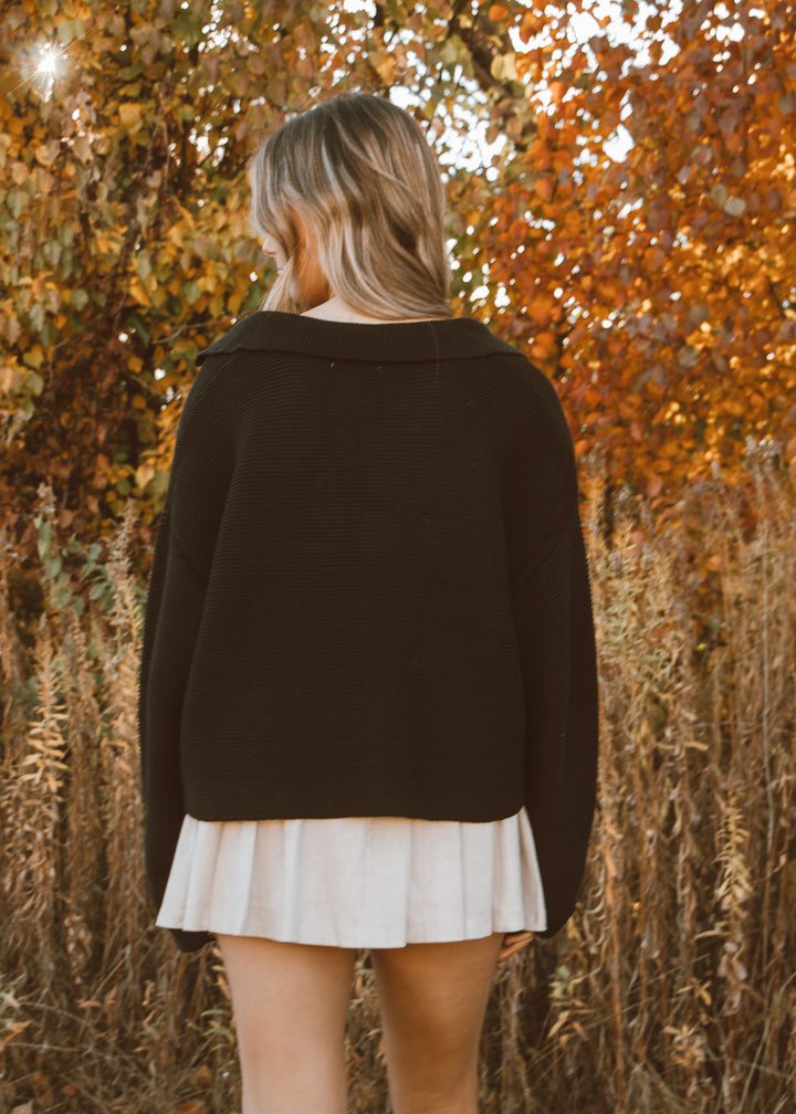 Person wearing a black sweater and white pleated skirt standing in front of autumn foliage.