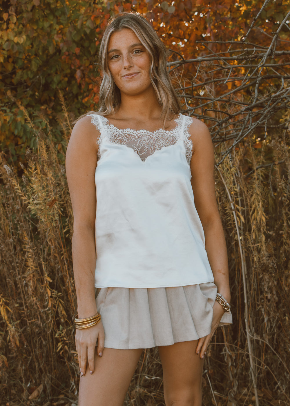 Woman wearing a white lace top and beige skirt standing in front of autumn foliage.