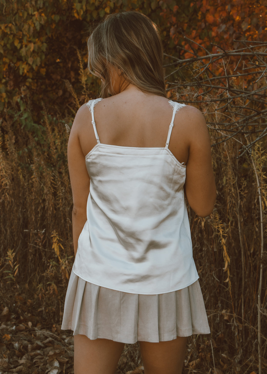 Woman wearing a white tank top and beige skirt standing in a natural setting with brown foliage.