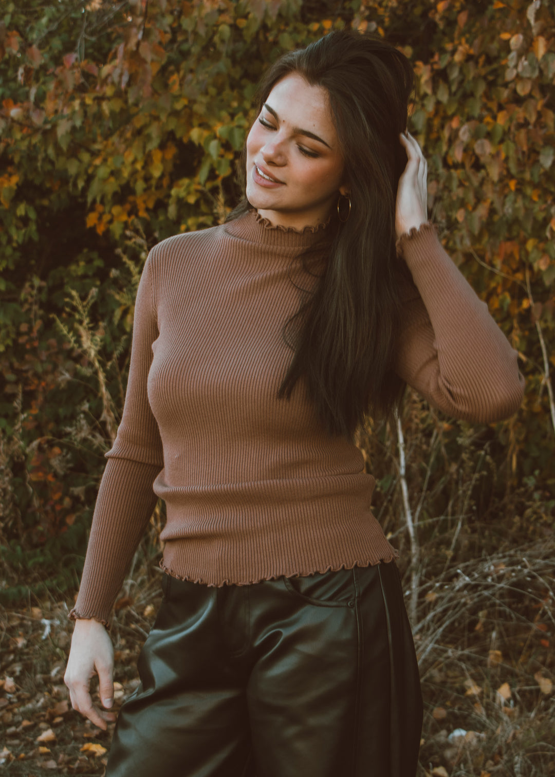 Woman in a brown turtleneck top and leather pants standing in a natural setting with trees and foliage.