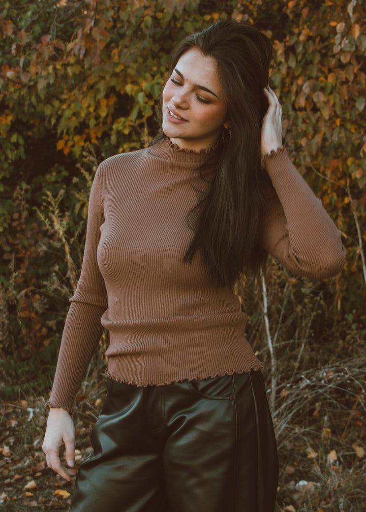 Woman in a brown turtleneck top and leather pants standing in a natural setting with trees and foliage.