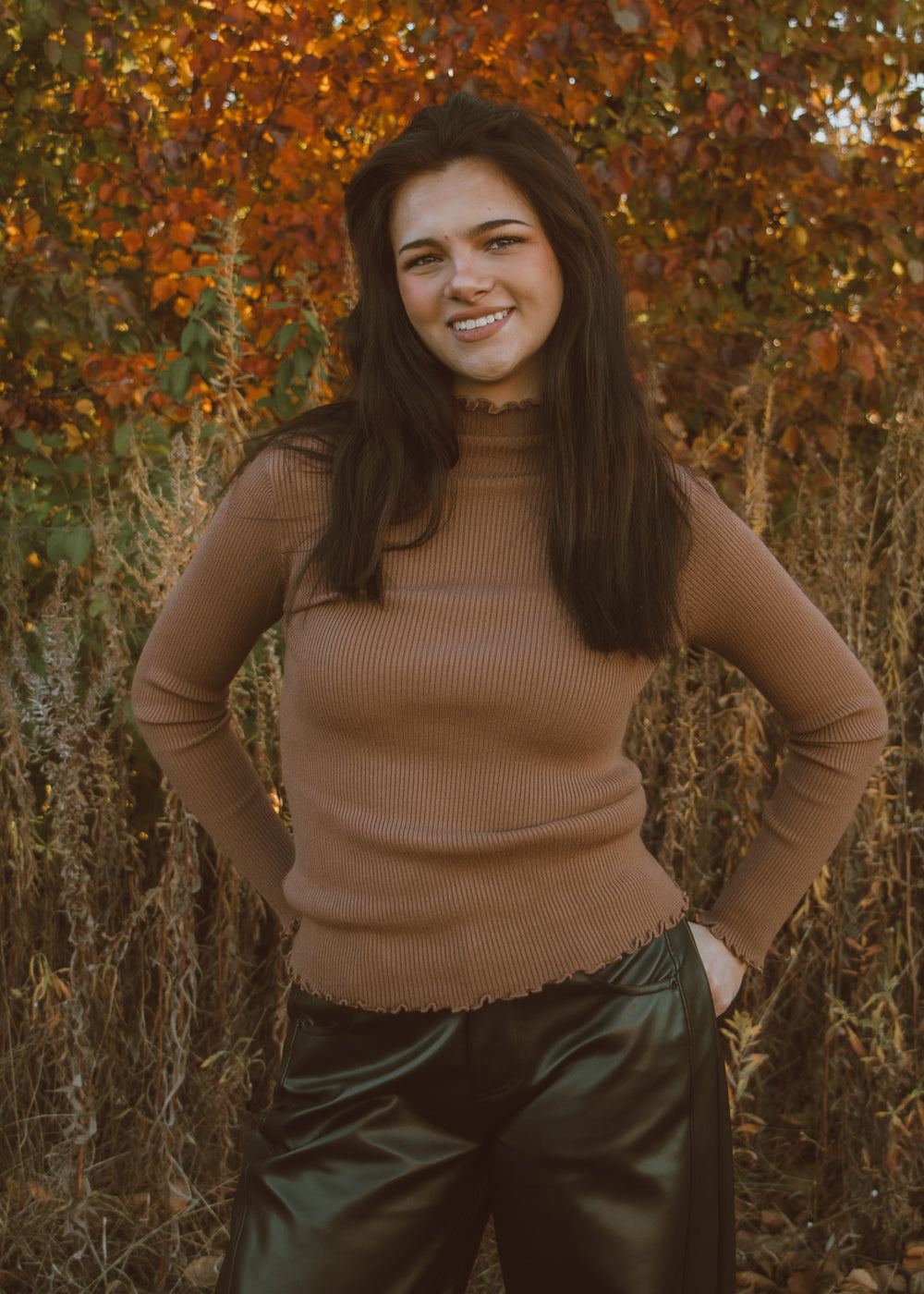 Woman wearing a brown ribbed top and leather pants standing in front of autumn foliage.