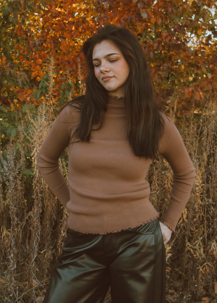 Woman in a brown ribbed turtleneck top and black pants standing against an autumnal background.