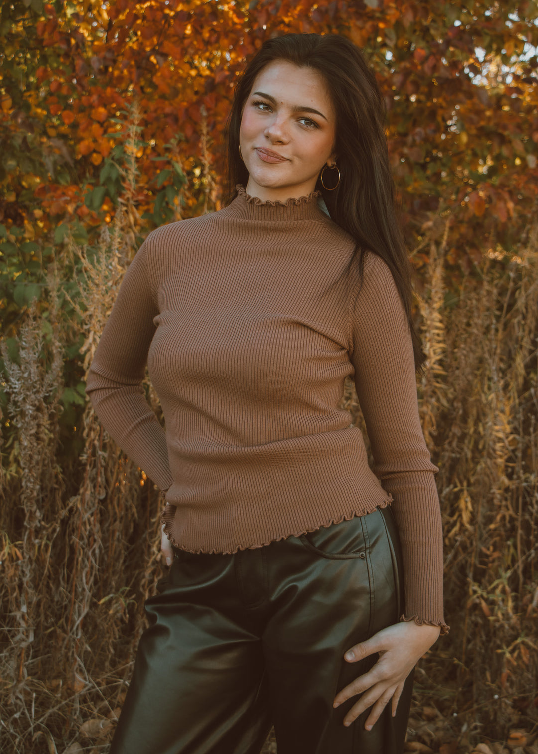 Woman wearing a brown turtleneck top and black leather pants standing in front of autumn foliage.