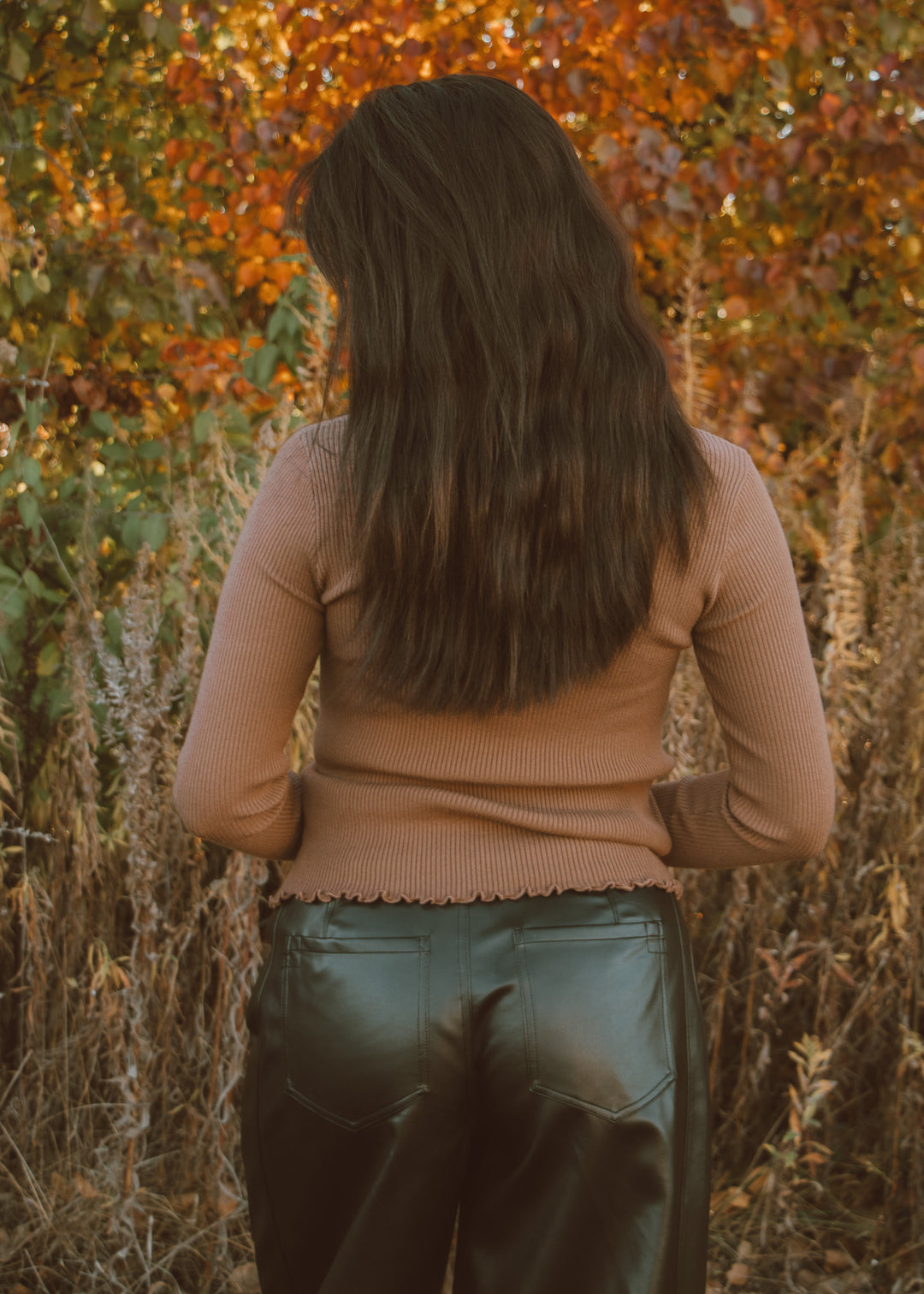 Person wearing a brown top and leather pants standing in front of autumn foliage.
