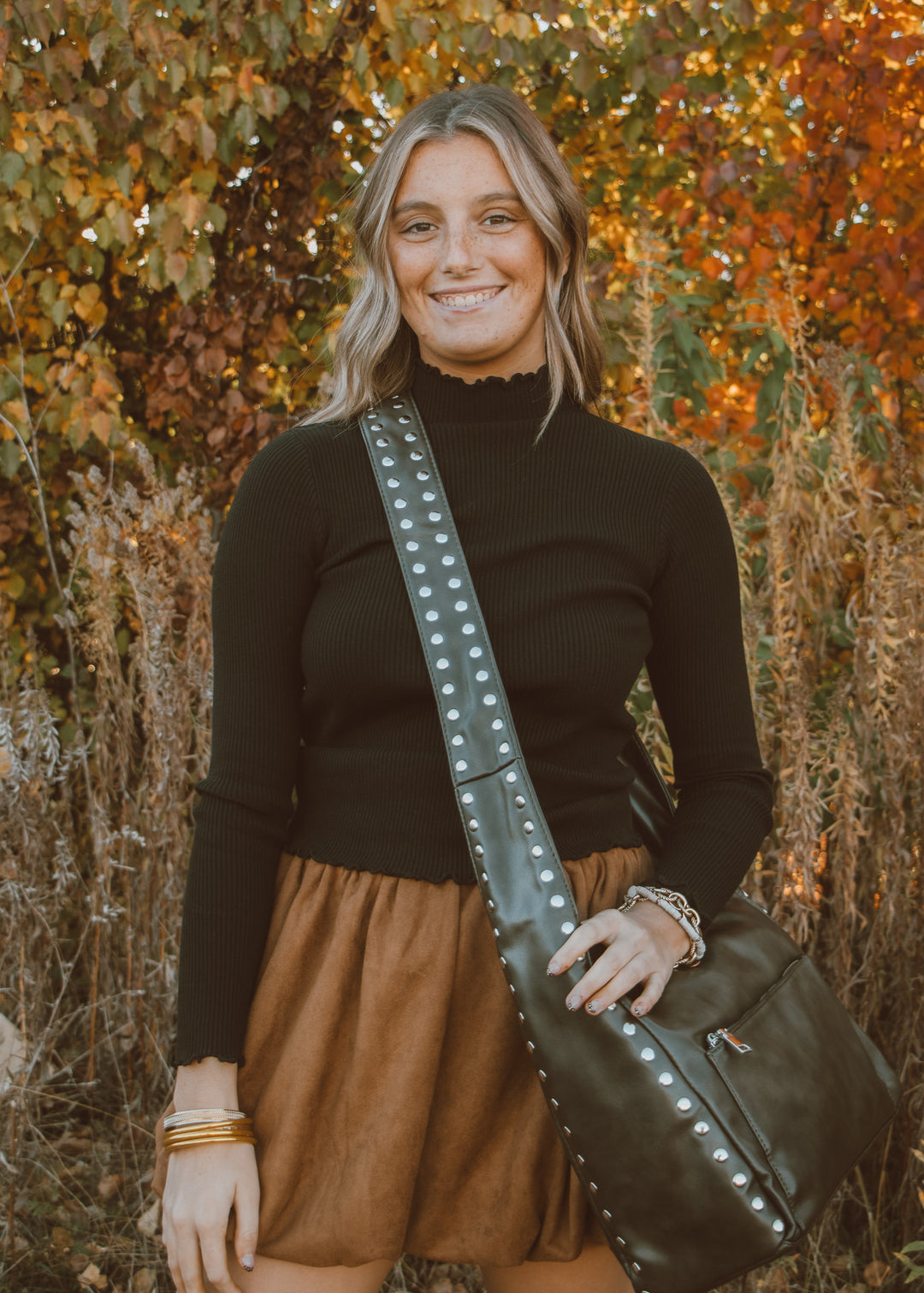 Woman holding a brown leather bag with studded details with a black top and bubble skirt in an autumn setting