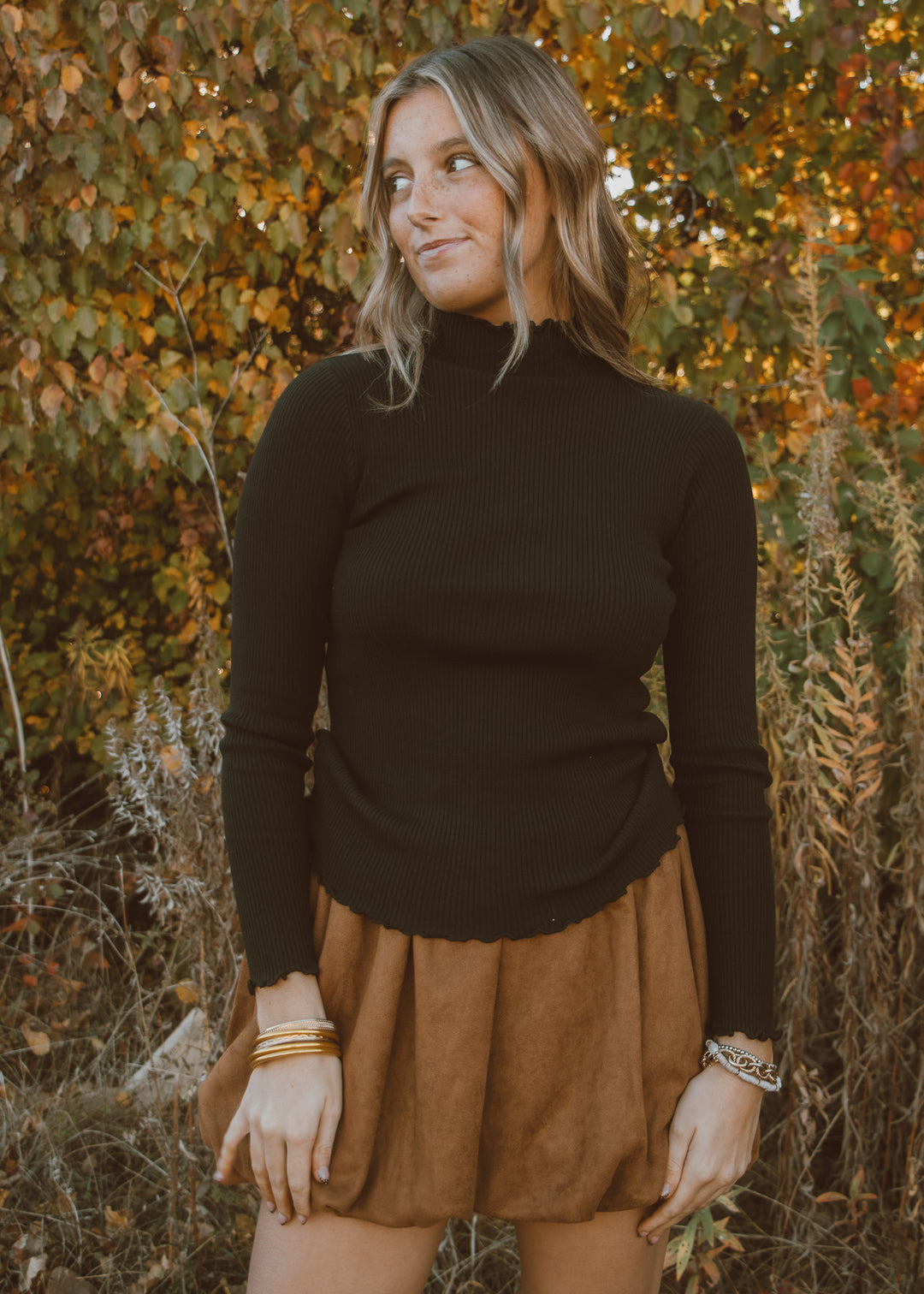 Woman wearing a black turtleneck and brown skirt standing in front of autumn foliage.
