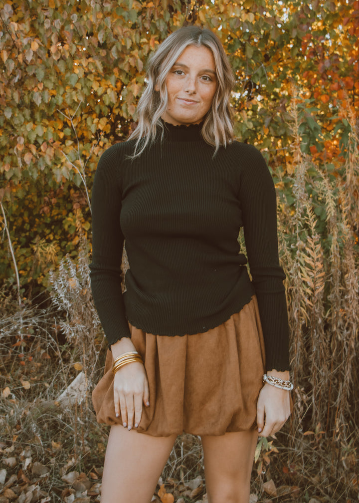 Woman wearing a black turtleneck and brown skirt standing in front of autumn foliage.