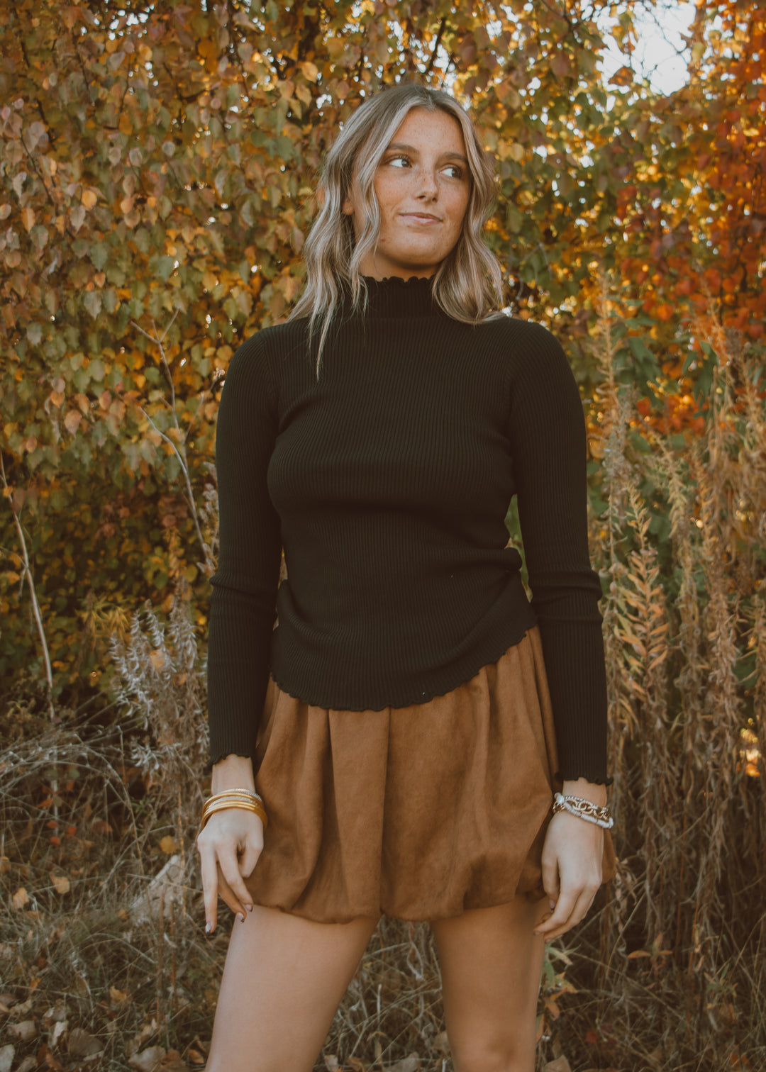 Woman wearing a black turtleneck long-sleeve top and brown skirt standing in an autumn setting.