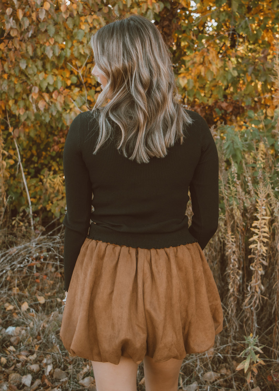 Person wearing a black top and brown skirt standing in an autumn setting with trees and dry leaves.