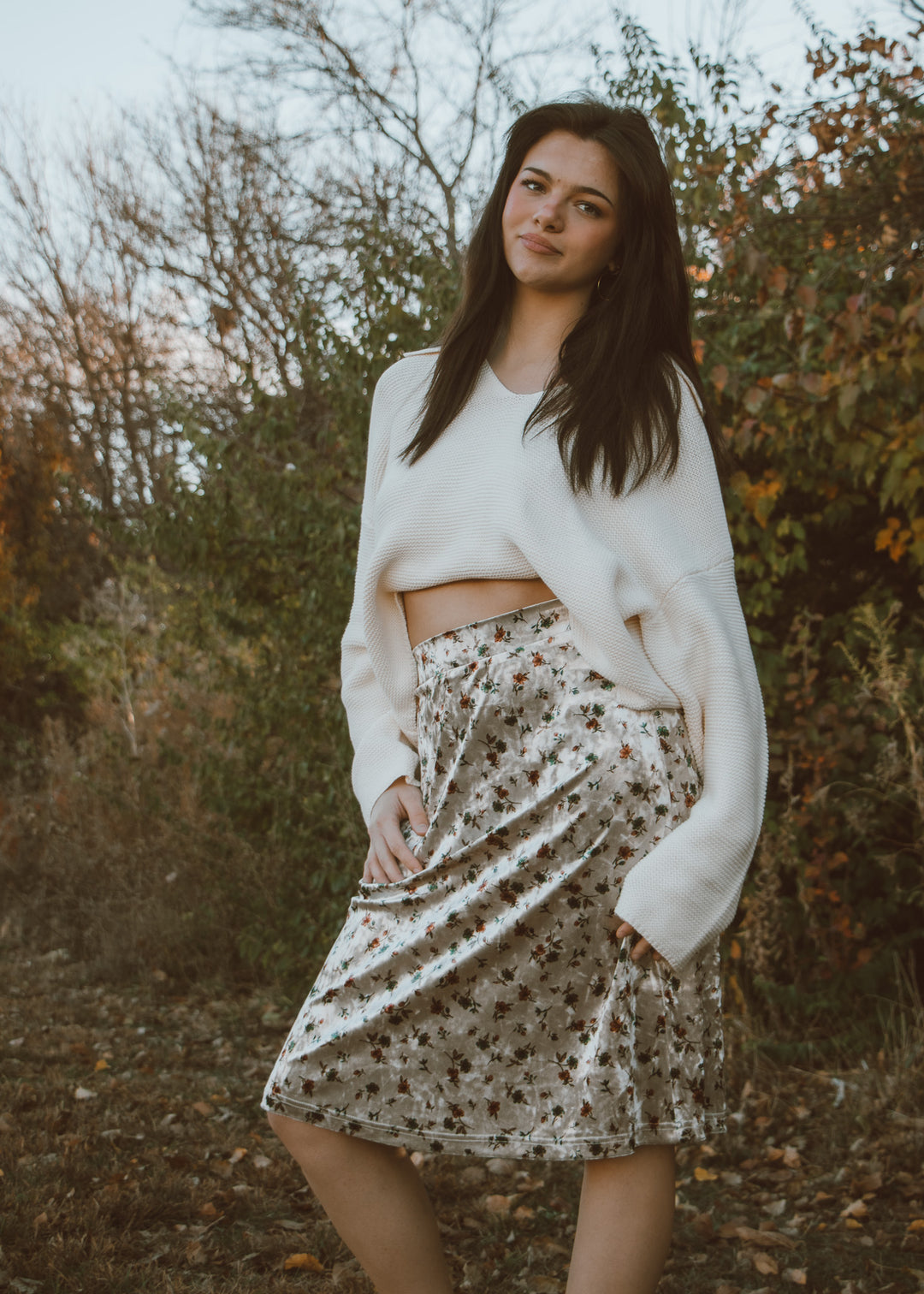 Woman wearing a white sweater and velvet floral skirt standing in a natural setting with trees and bushes.