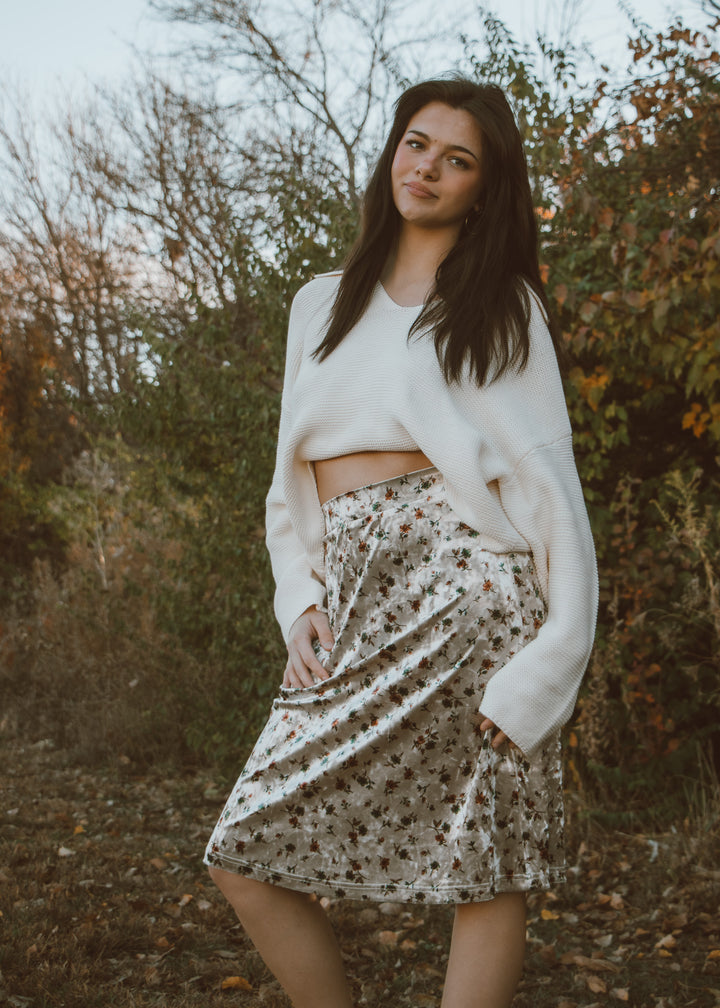 Woman wearing a white sweater and velvet floral skirt standing in a natural setting with trees and bushes.