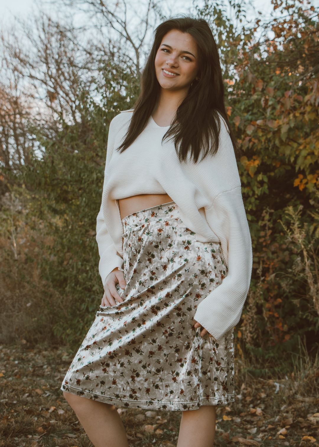 Woman wearing a white sweater and patterned skirt standing outdoors with trees in the background